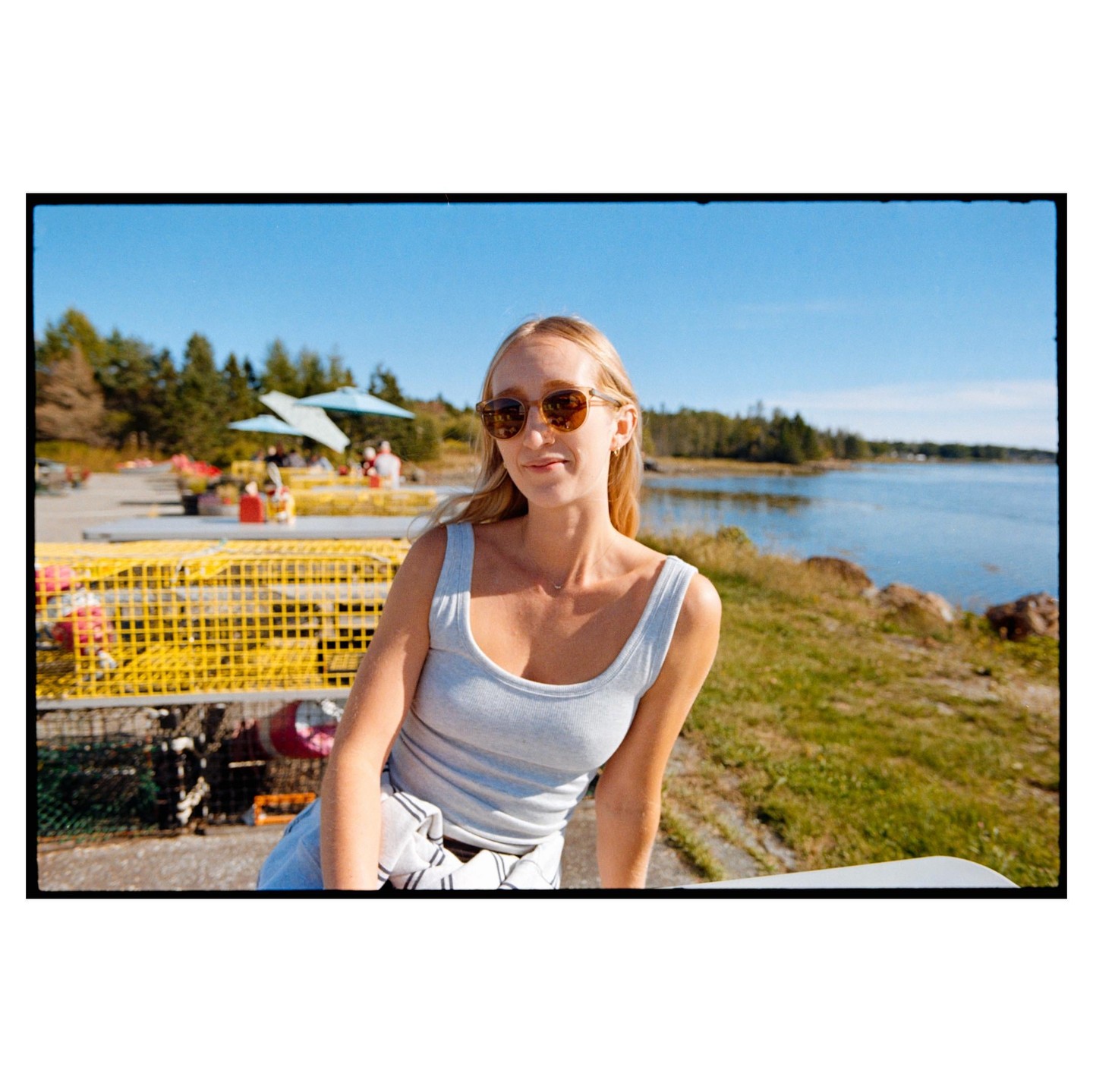 A woman wearing sunglasses and a tank top smiles while sitting outdoors near a body of water with lobster traps in the background.
