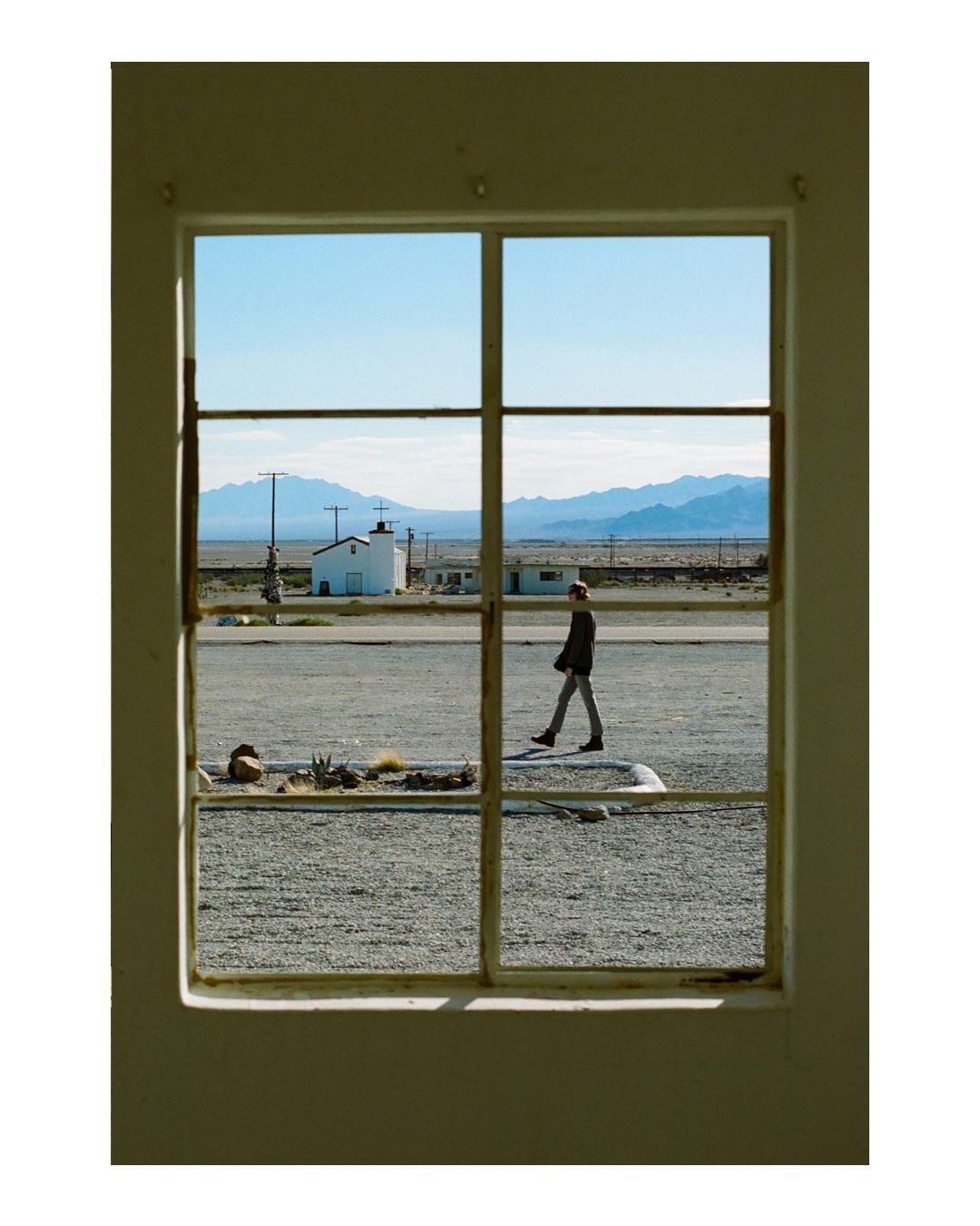 A man walks across a gravel lot towards a small white building with mountains in the background, viewed through a window frame.
