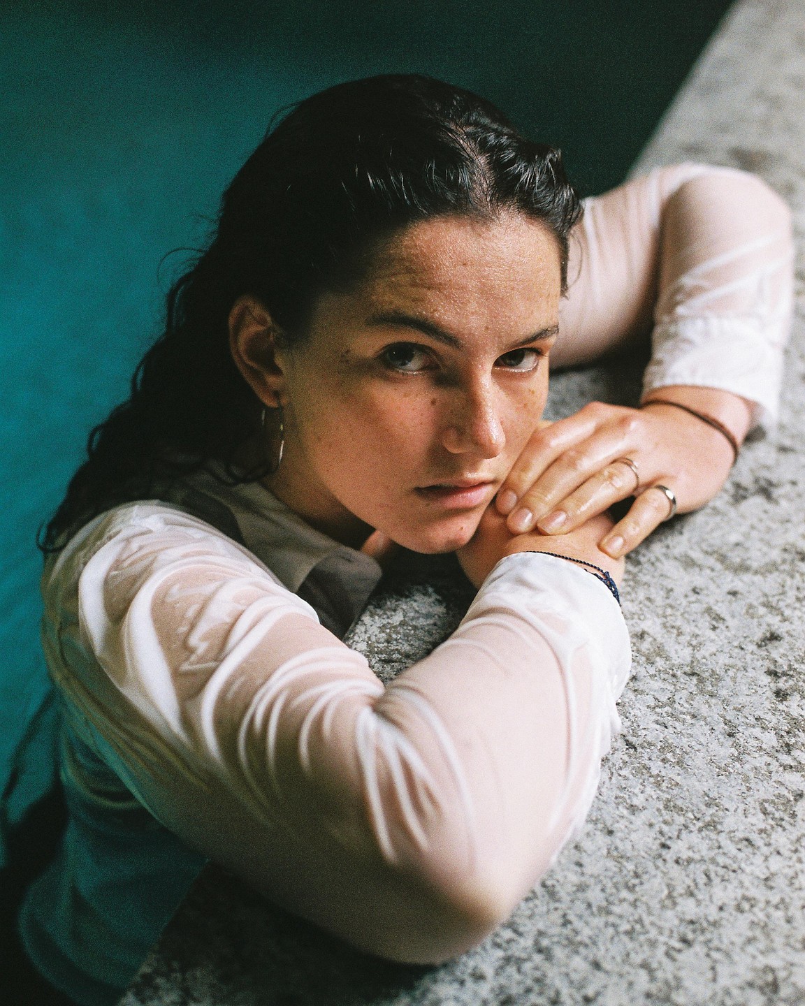 A woman with wet hair and a wet white shirt leans on a stone ledge, looking directly at the viewer.