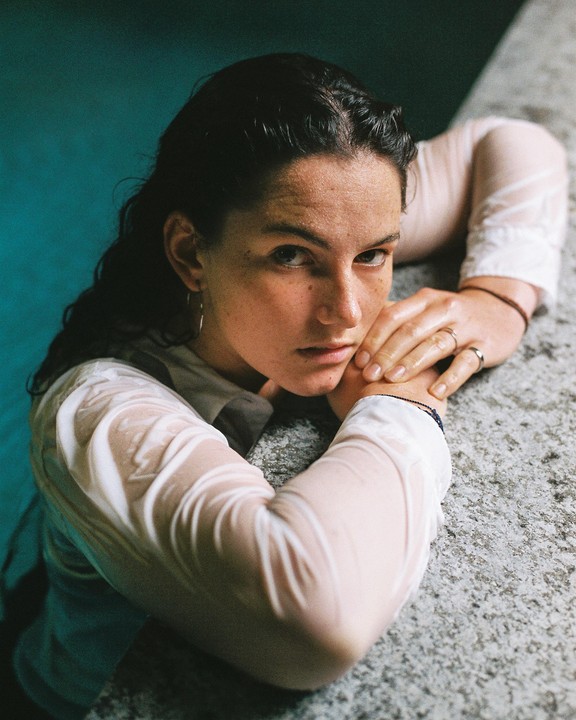 A woman with wet hair and a wet white shirt leans on a stone ledge, looking directly at the viewer.