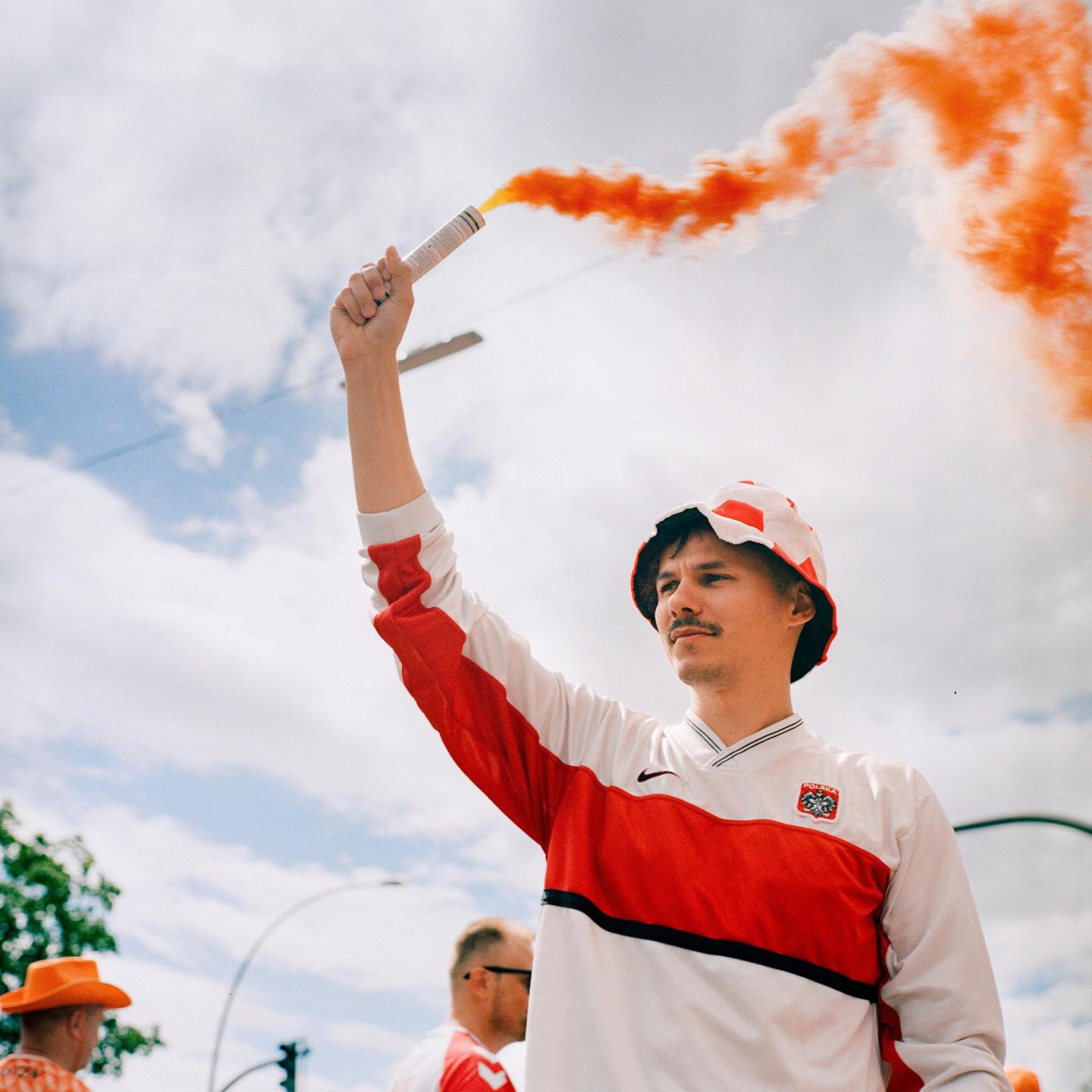 A man in a red and white shirt and bucket hat holds an orange smoke flare.