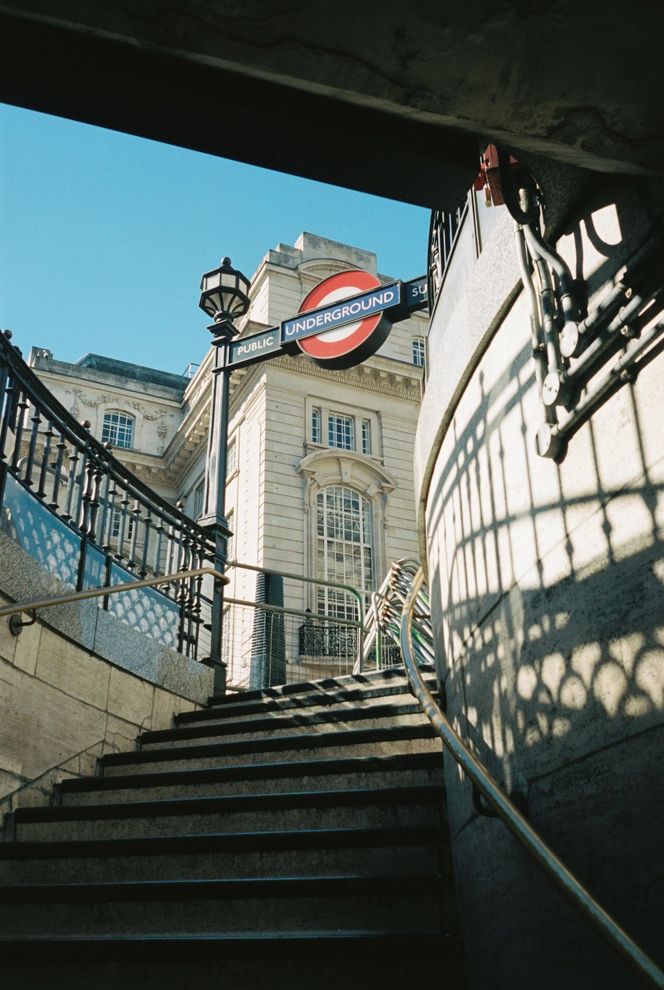 A low-angle view of a London Underground sign and a building, seen from the bottom of a staircase.