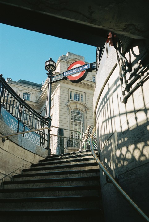 A low-angle view of a London Underground sign and a building, seen from the bottom of a staircase.