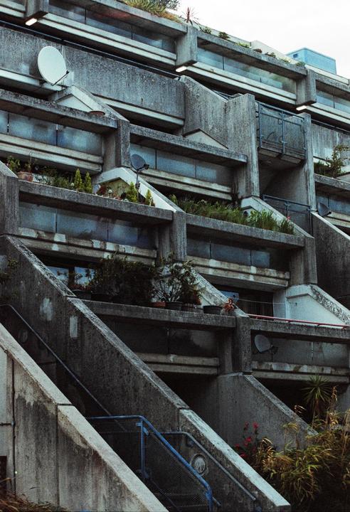 A brutalist concrete building with plants on balconies and satellite dishes on its facade is shown from a low angle.