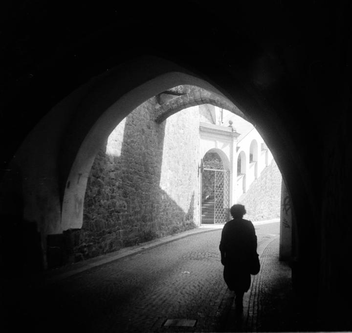 A person walks through a dark archway towards a sunlit street with stone walls and a gate.