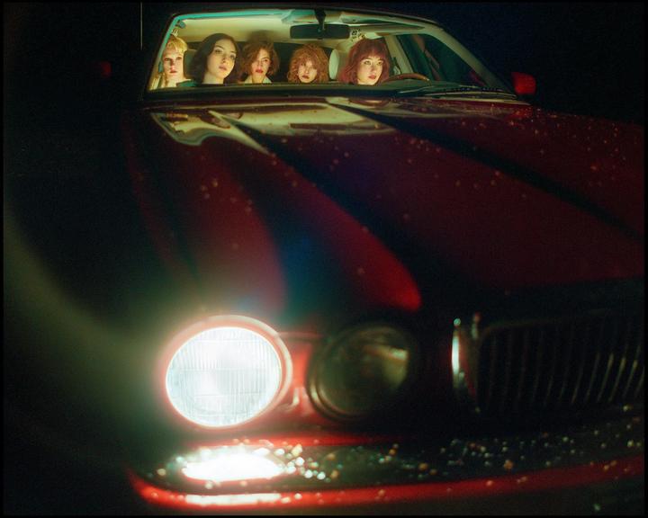 Four women are visible through the windshield of a red car at night, with one headlight shining brightly.