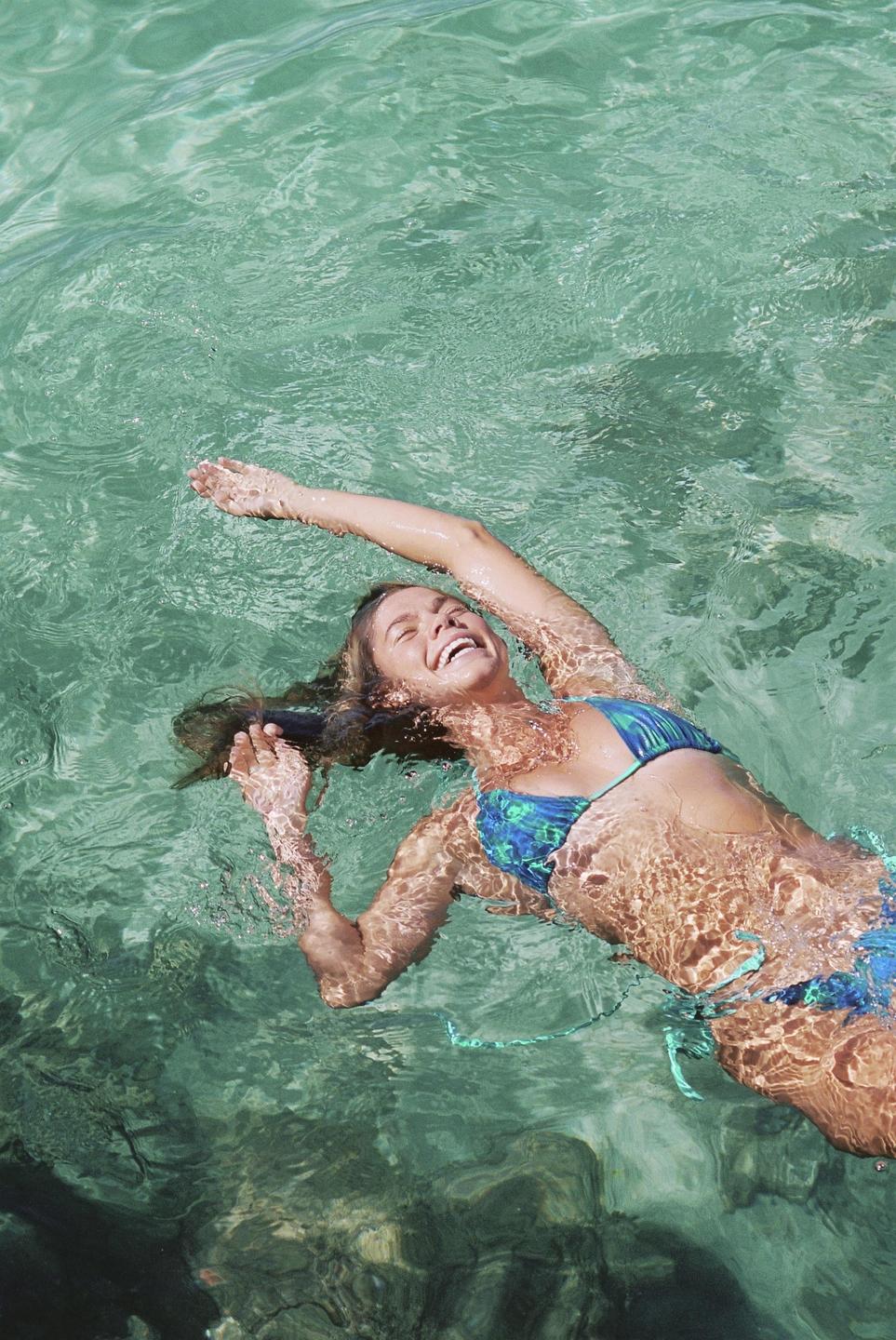 A woman in a blue bikini floats on her back in clear green water, laughing with her arms outstretched.