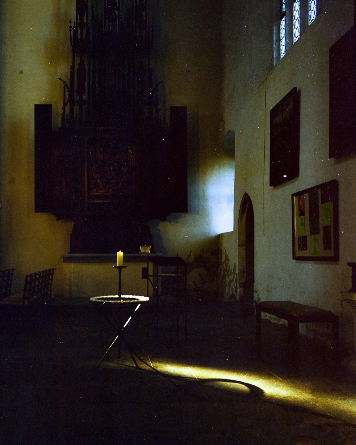 A single candle burns on a stand in a dark room with light coming through a window.