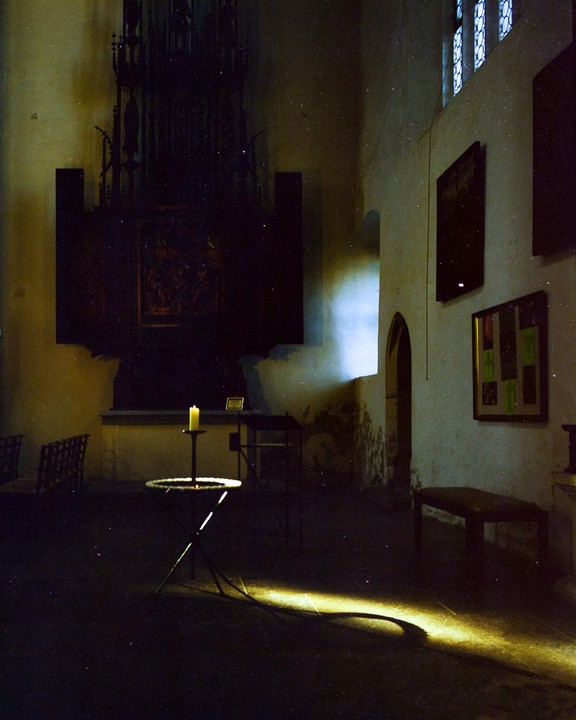 A single candle burns on a stand in a dark room with light coming through a window.
