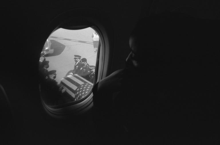 A person looks out an airplane window at a flag-draped coffin being carried by uniformed men on an airfield.