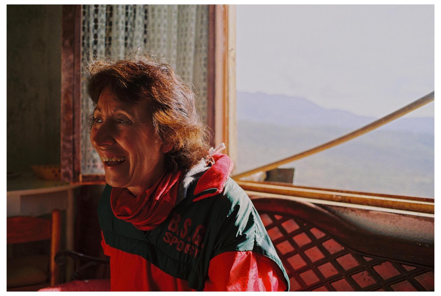 A woman with red hair smiles while sitting by a window overlooking mountains.