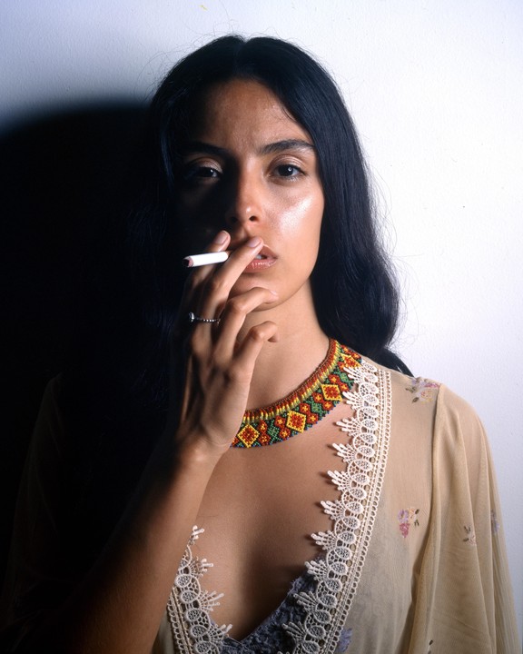 A woman with long dark hair holds a cigarette to her lips, wearing a colorful beaded necklace and a sheer top with lace trim.