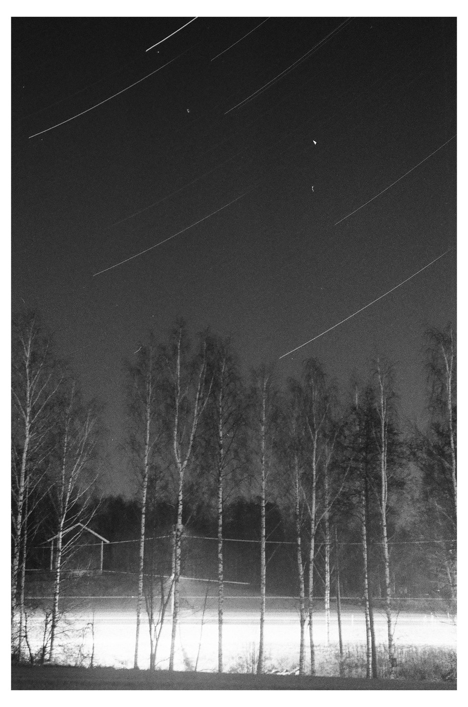 Star trails are visible above a line of trees and a small building next to a road.