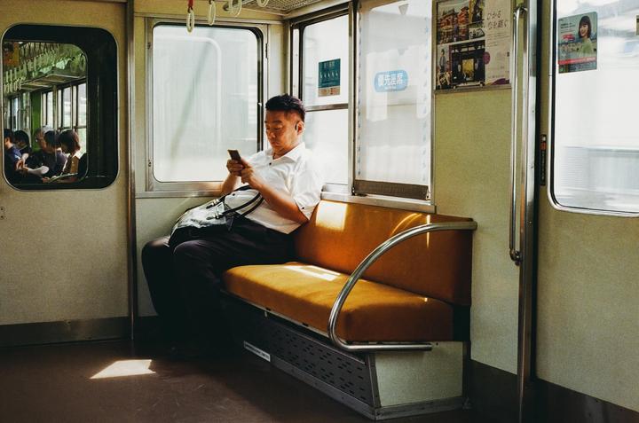 A man sits on a train bench looking at his phone, with other passengers visible in the reflection of a window.