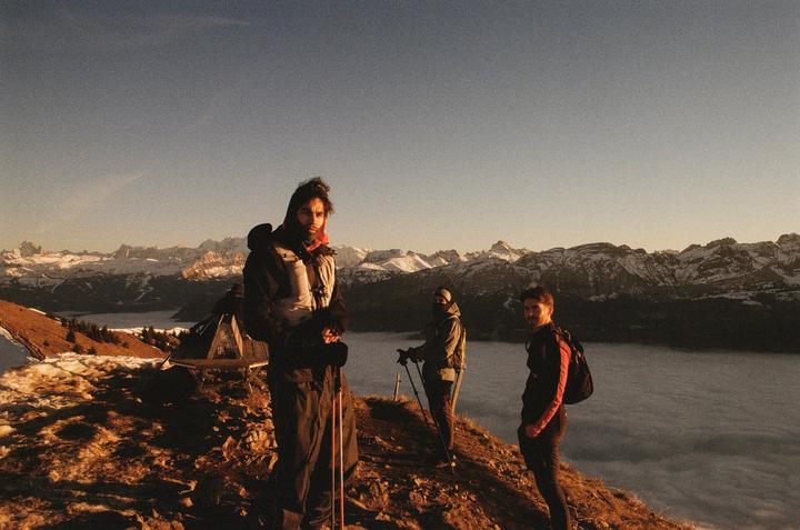 Three men stand on a mountain ridge overlooking a sea of clouds with snow-capped mountains in the distance.