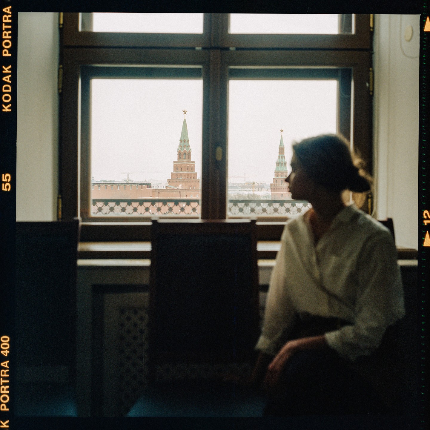 A woman sits in a room, looking out a window at two distant towers with red stars on top.