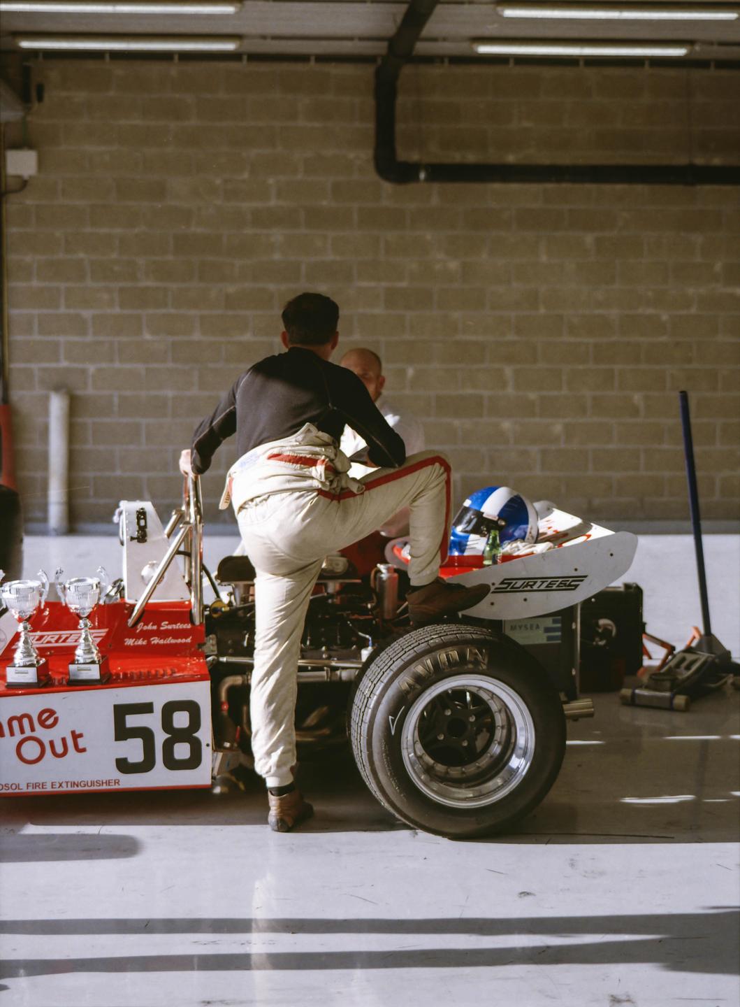 A man in a racing suit stands next to a red race car with another man sitting in the background.