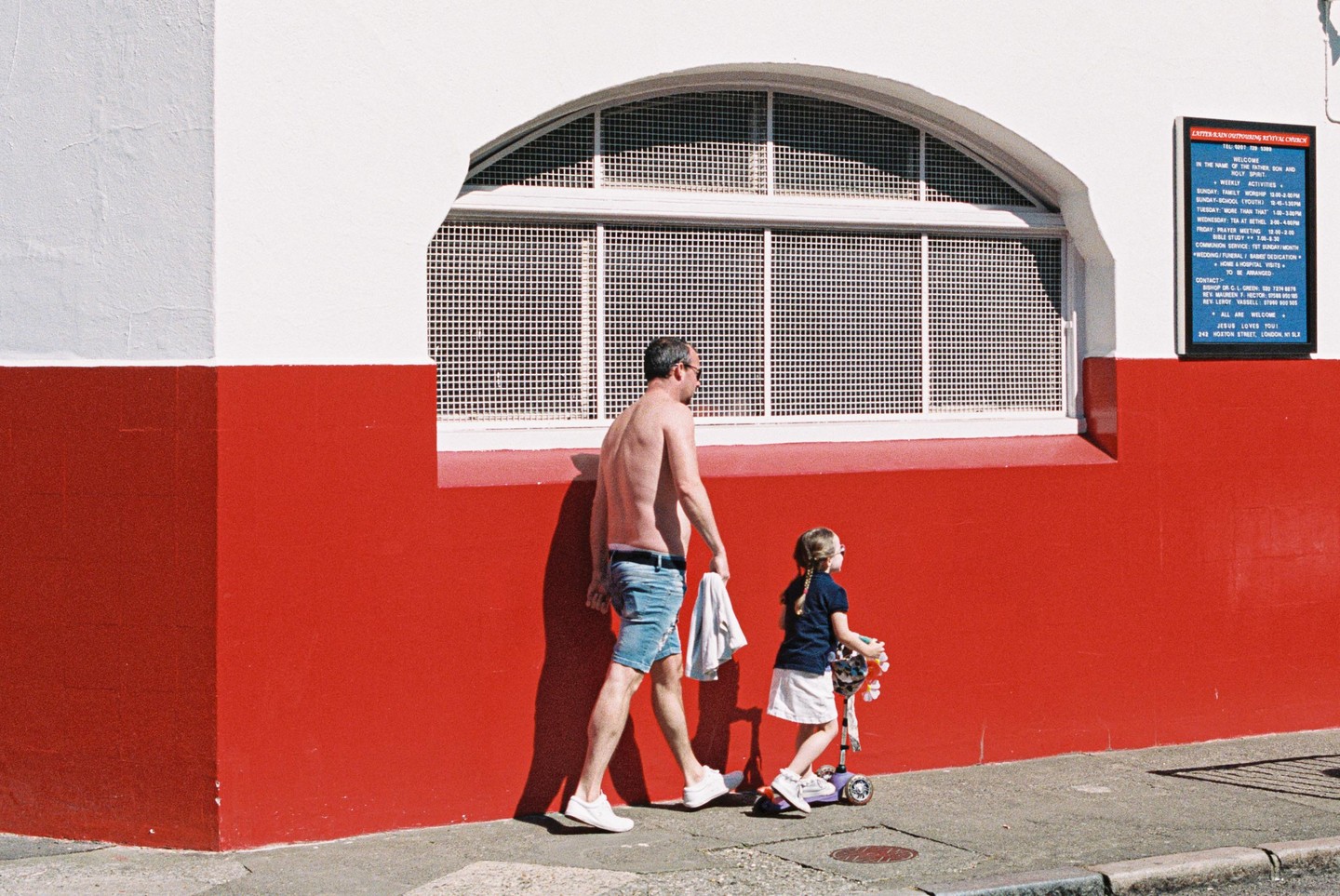 A man and a girl with a scooter walk past a red and white building with an arched window.