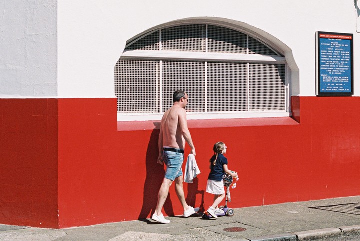 A man and a girl with a scooter walk past a red and white building with an arched window.