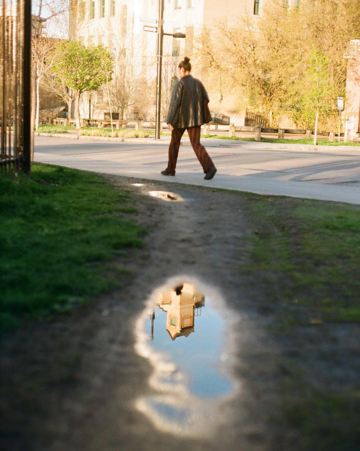 A person walks on a sidewalk next to a street with a reflection of a building and sky in a puddle in the foreground.