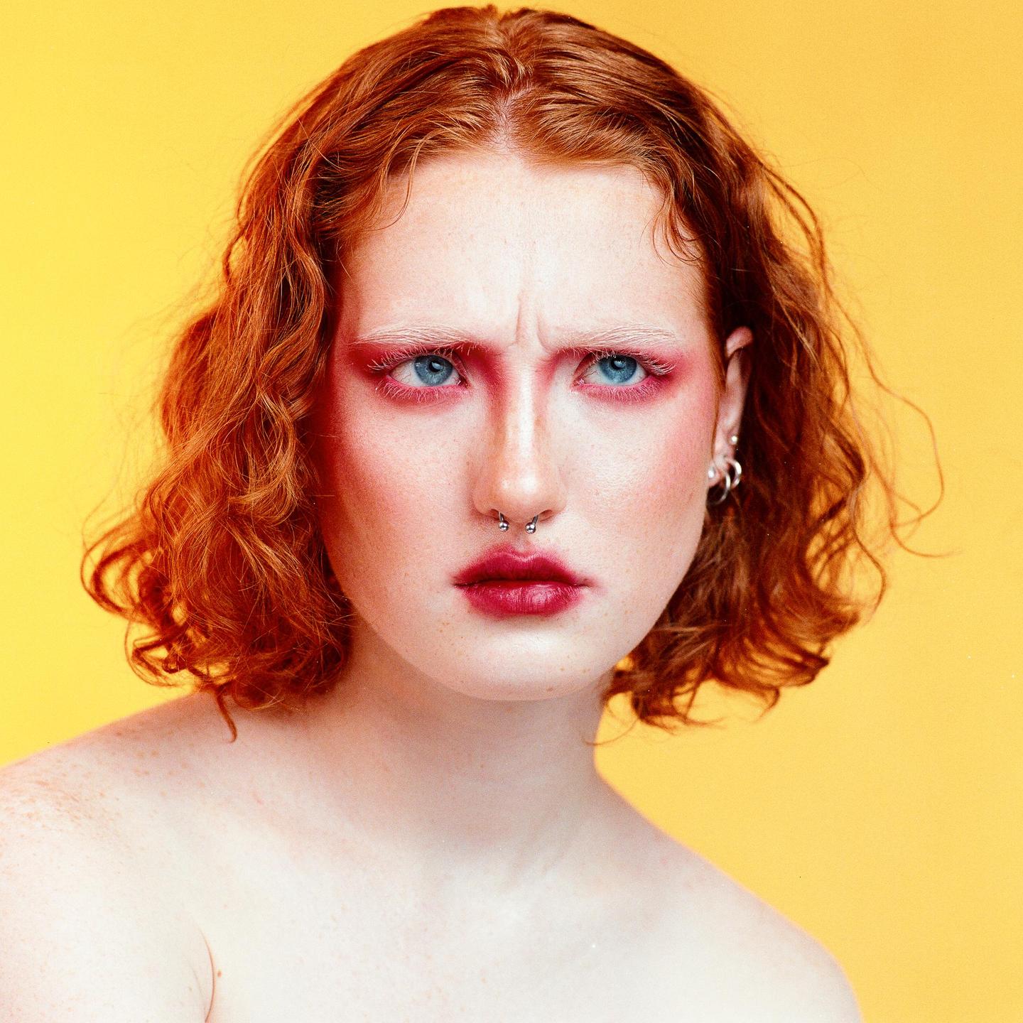 A woman with red hair and red eye makeup looks directly at the viewer with a furrowed brow against a yellow background.