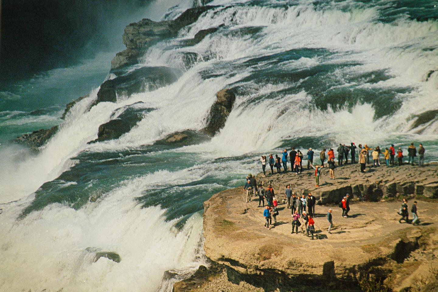 A large group of people stand on a rocky outcrop next to a powerful waterfall.