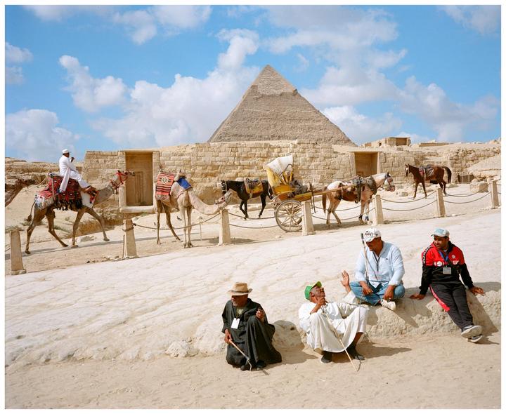 Several men are gathered in a desert landscape with camels, horses, and a pyramid in the background.