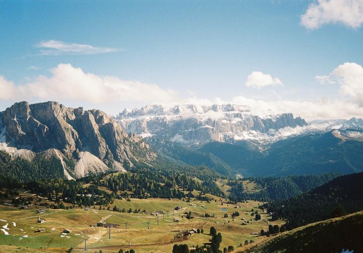 A vast mountain landscape features a valley with scattered buildings and trees, leading up to a range of rocky peaks, some of which are snow-capped.