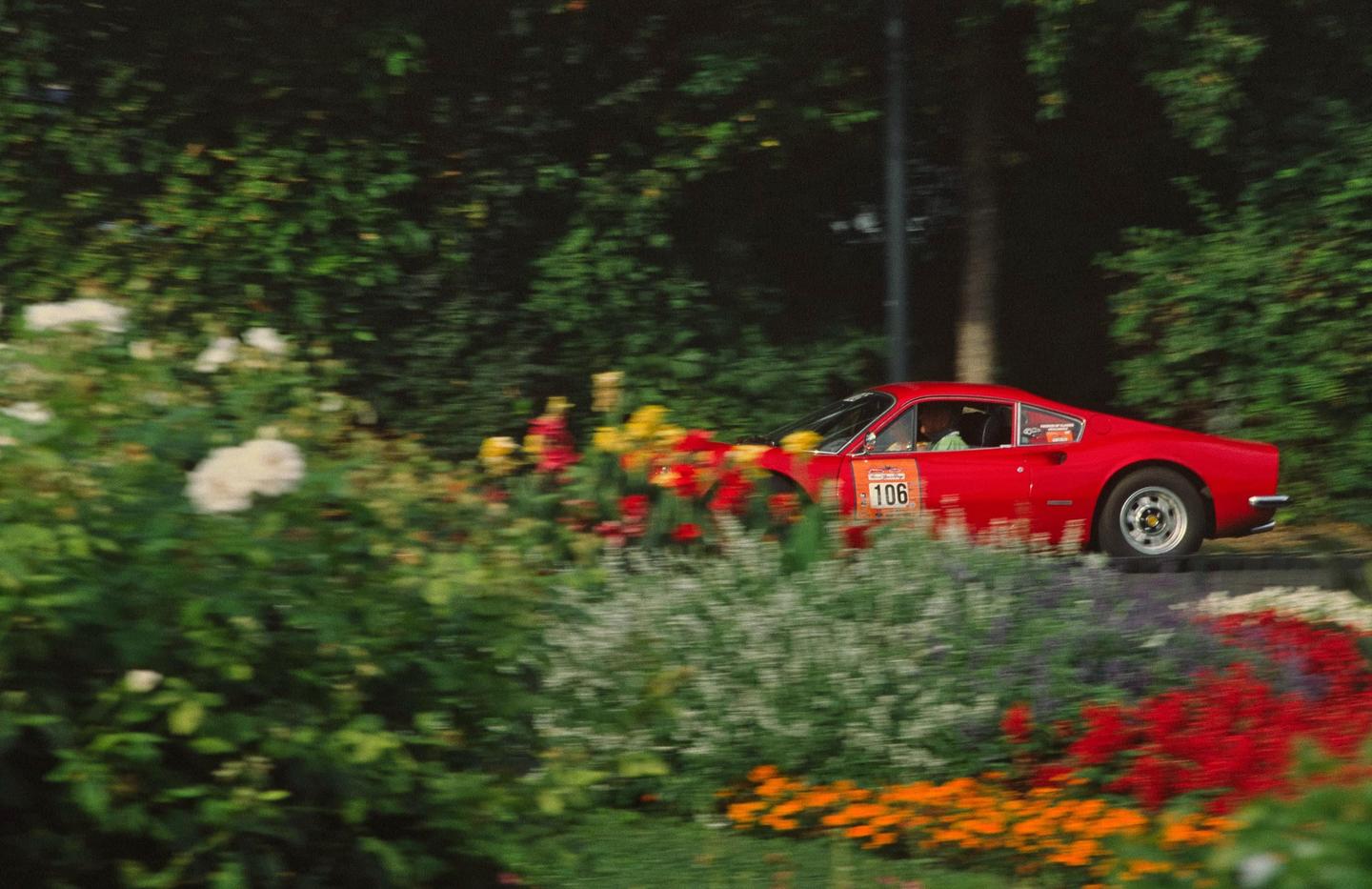 A red car with the number 106 on its side drives past a garden with colorful flowers and green bushes.
