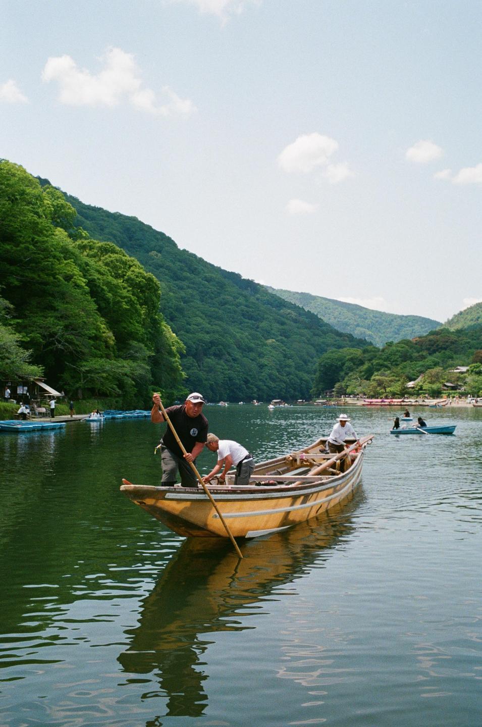 Three men are in a wooden boat on a river surrounded by lush green mountains.
