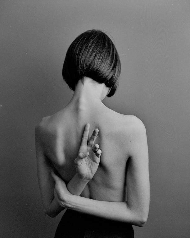 A woman with short hair stands with her back to the viewer, holding her hands behind her back with two fingers raised in a peace sign.