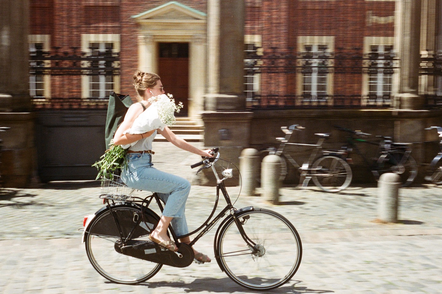 A woman rides a bicycle on a cobblestone street, holding a bouquet of white flowers and carrying a green bag.
