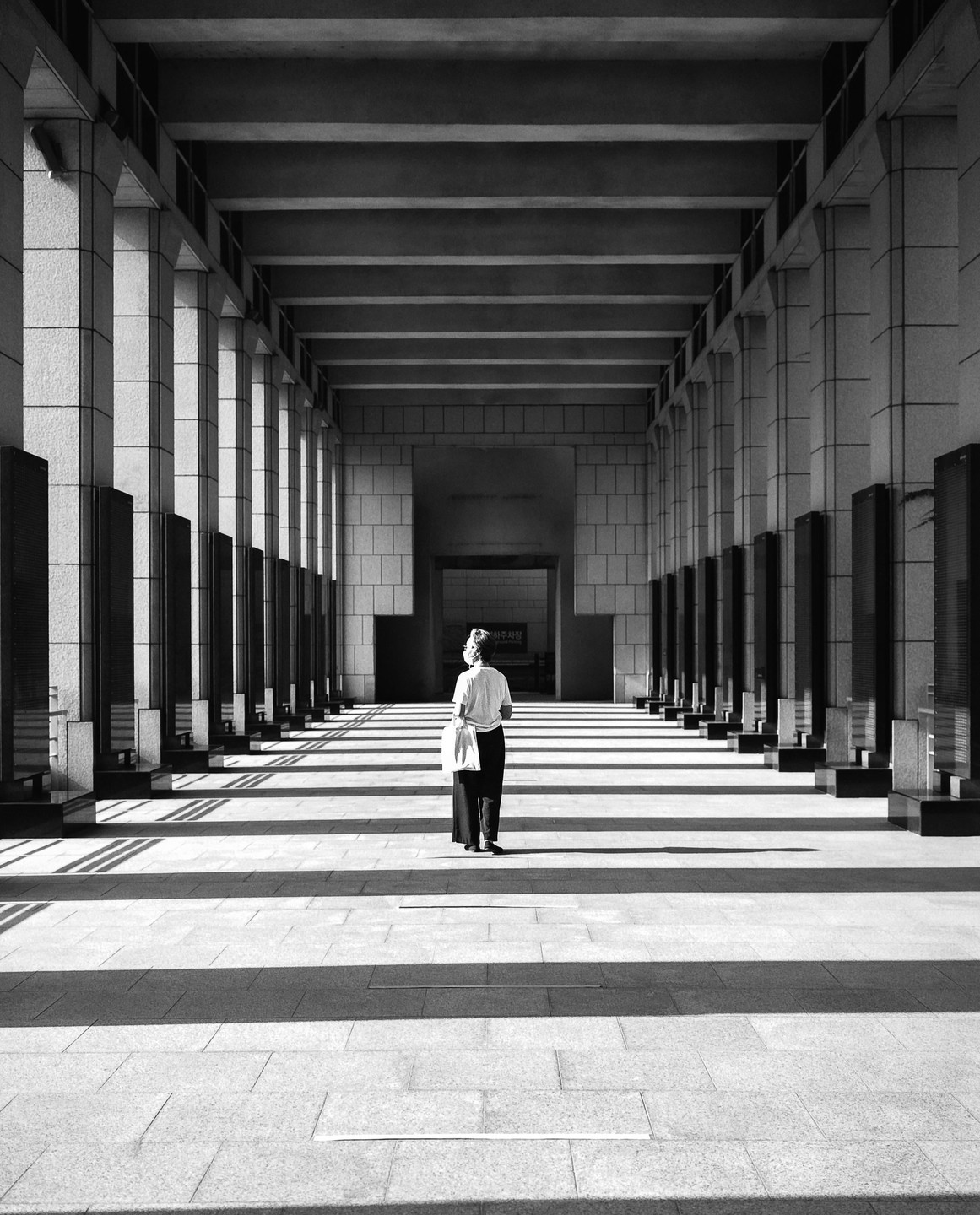 A woman stands in a long, columned hallway with strong shadows stretching across the floor.