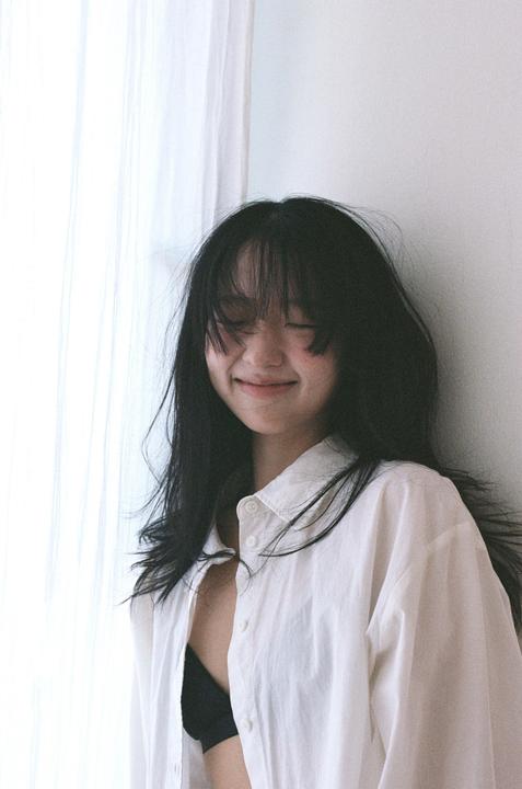 A woman with long dark hair and bangs smiles with her eyes closed, wearing a white shirt and black bra.