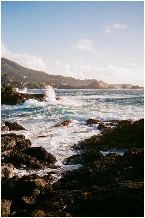 Waves crash against a rocky coastline with a distant mountainous landscape under a partly cloudy sky.