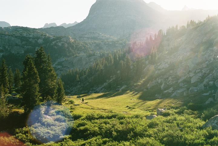 A person walks along a path through a green valley surrounded by mountains and trees.