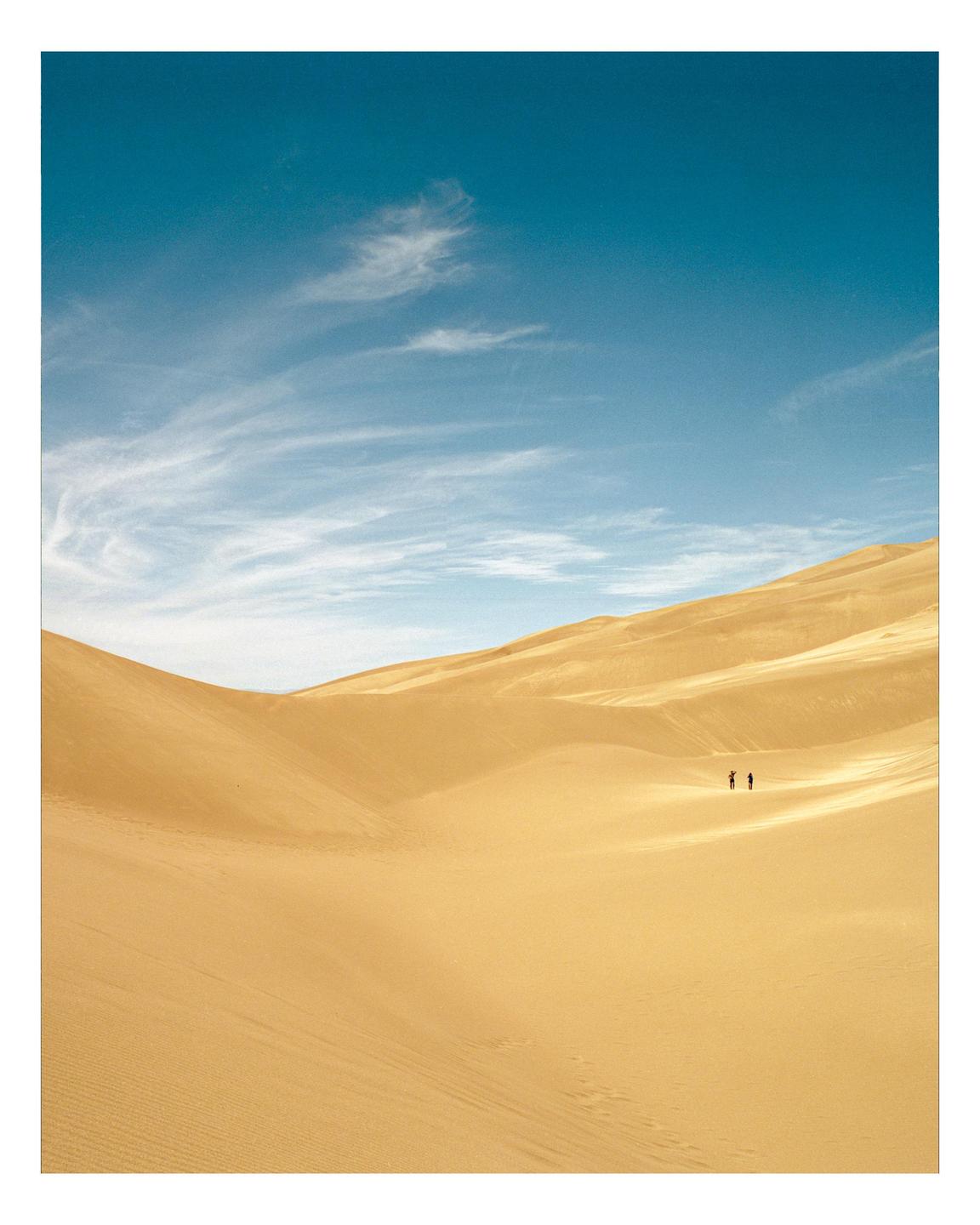 Two people stand in the distance on a vast sand dune under a blue sky with wispy clouds.