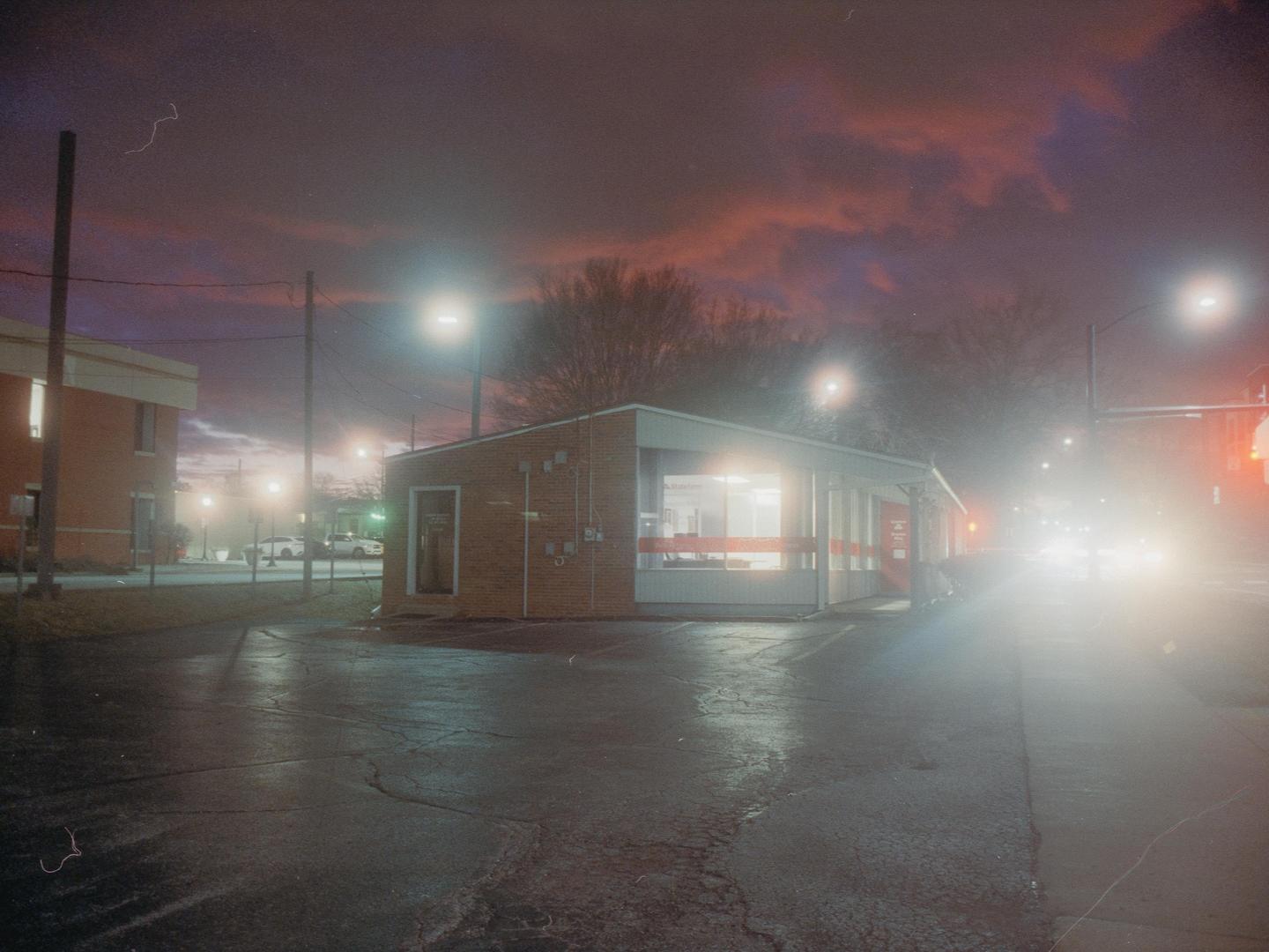 A brick building with large windows is illuminated at night under a dramatic sky, with bright streetlights and car headlights creating a hazy glow.