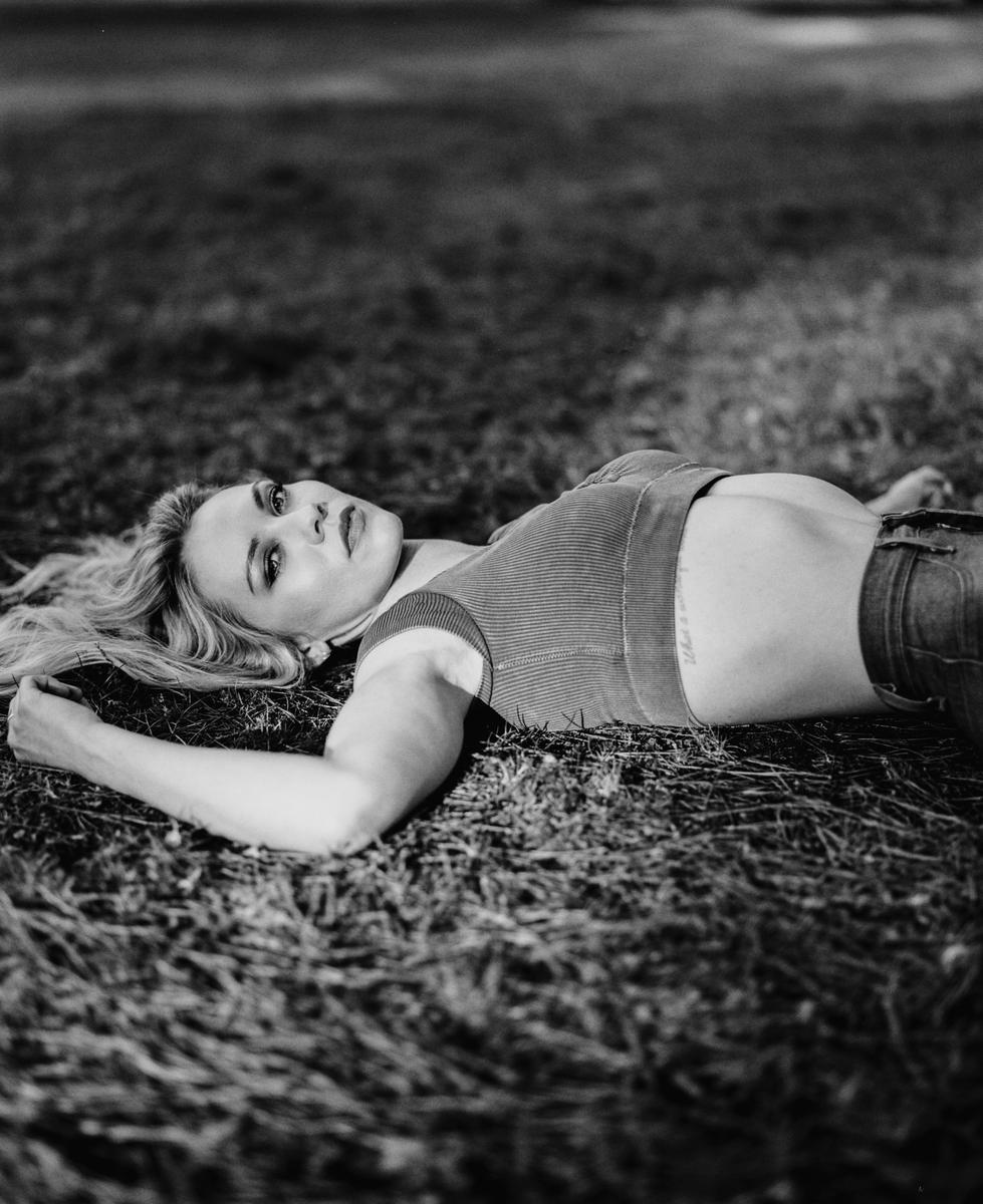 A woman with long hair lies on her back in the grass, looking up at the viewer.