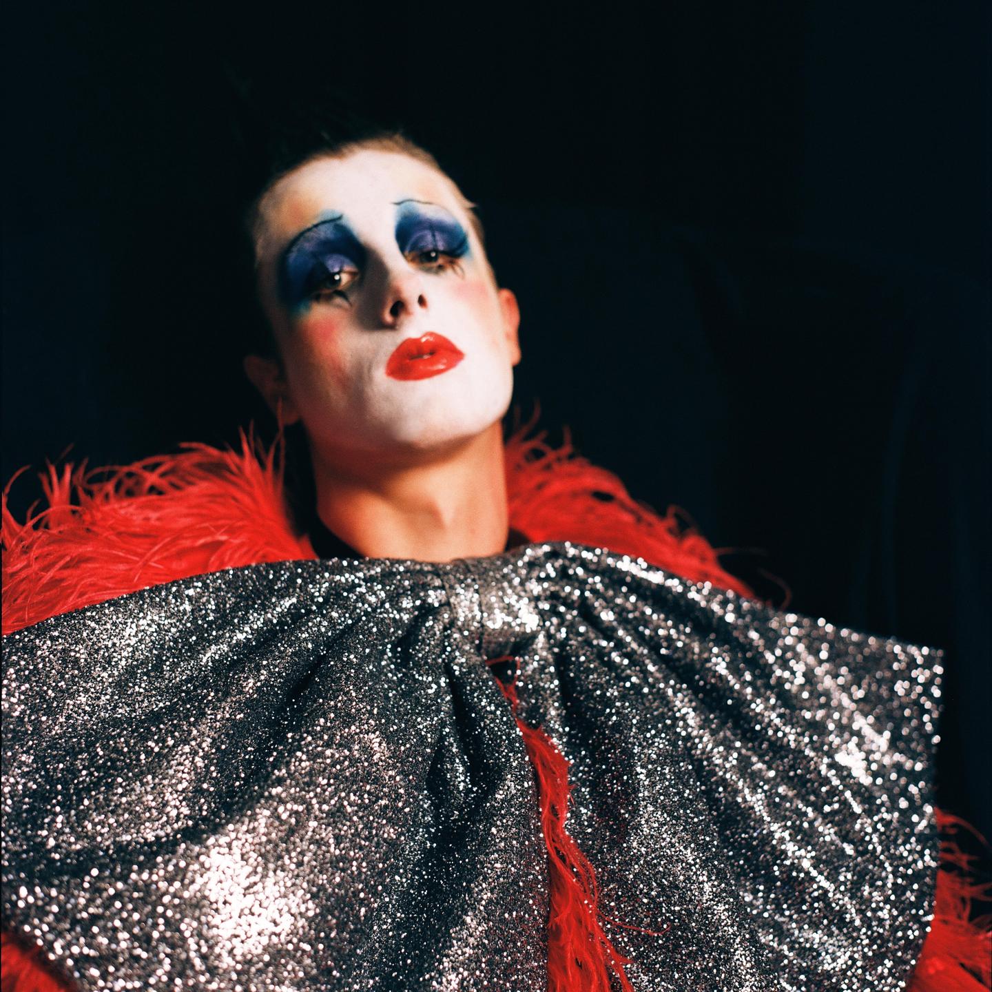 A person with clown makeup and a sequined garment with red feathers looks up.