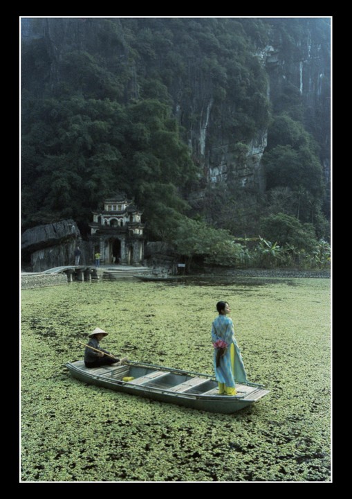 A woman in a traditional dress stands in a boat on a lake covered in green plants, with a temple and mountains in the background.