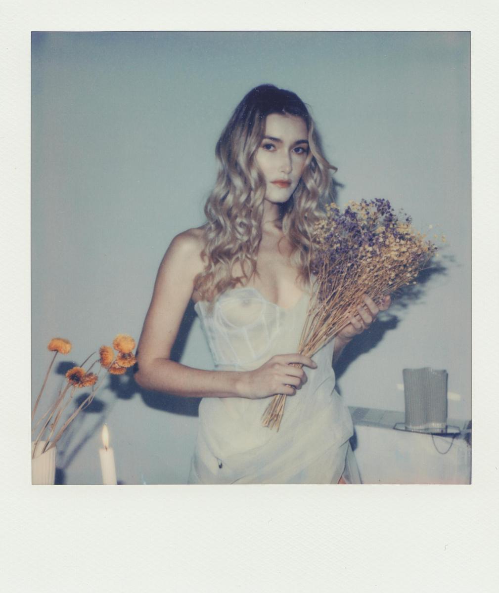 A woman with long, wavy hair holds a bouquet of dried flowers while standing next to a lit candle and other dried flowers.