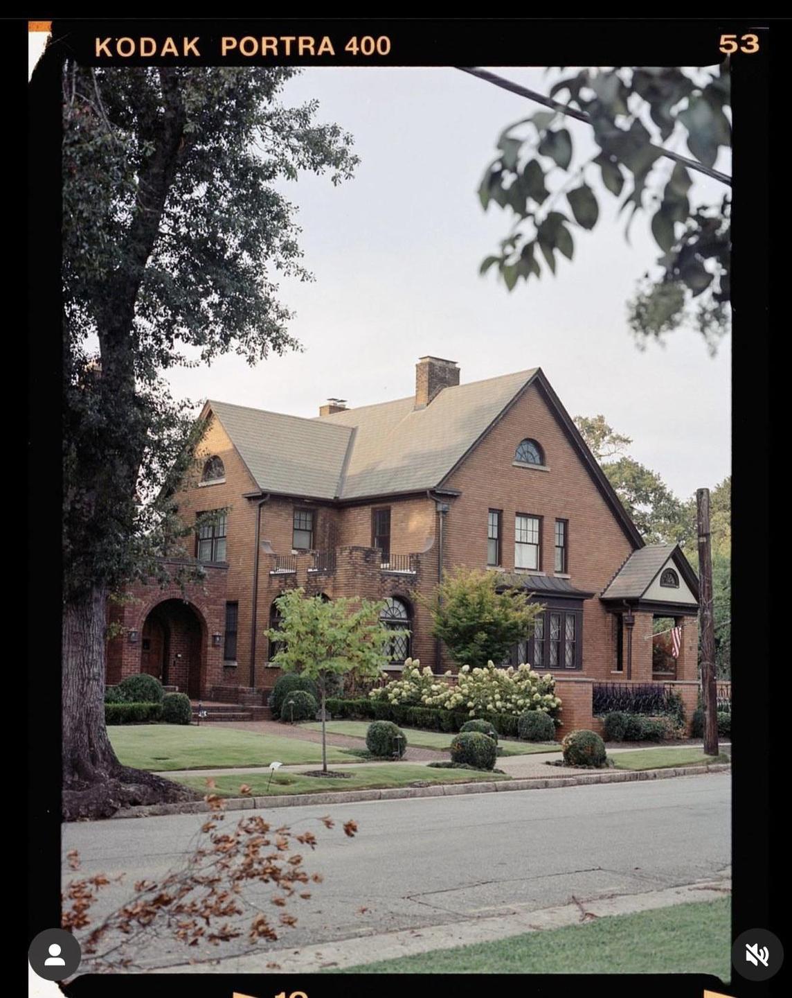 A large brick house with a green roof stands behind a manicured lawn and a street.