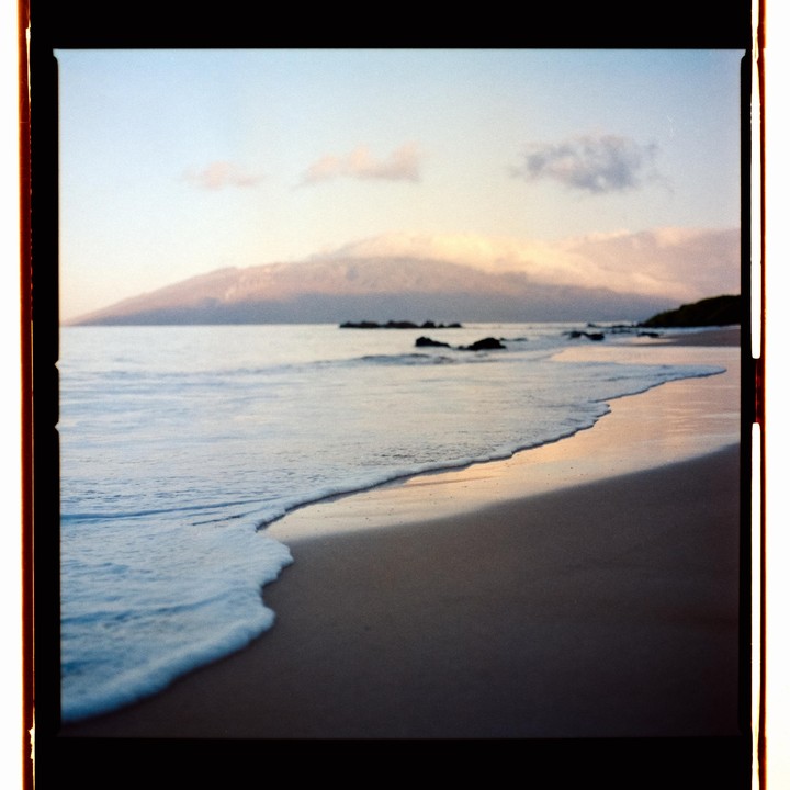 Waves gently wash onto a sandy beach with a mountain in the background under a cloudy sky.