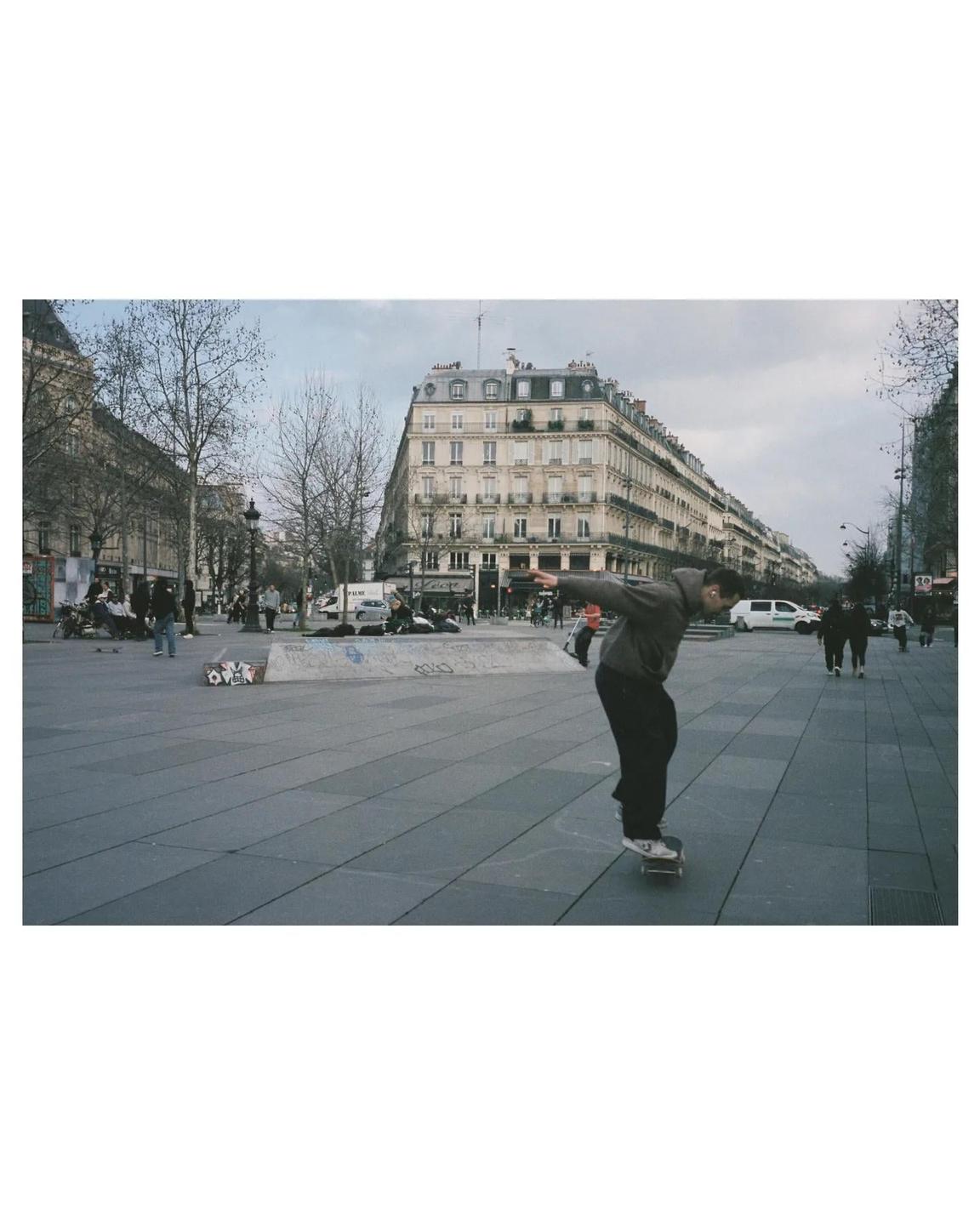A man on a skateboard balances in a city square with buildings and trees in the background.