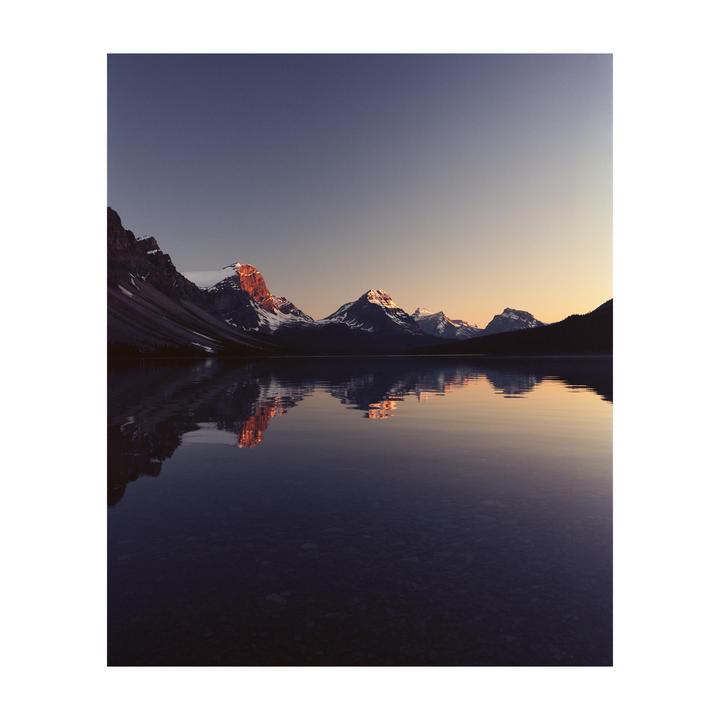 Snow-capped mountains are reflected in a calm lake under a clear sky at dusk.