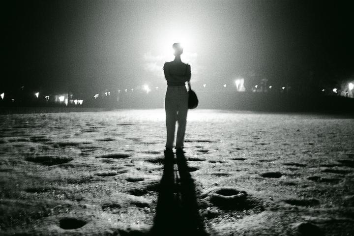 A person stands backlit by a bright light in a dark, textured field with distant lights.