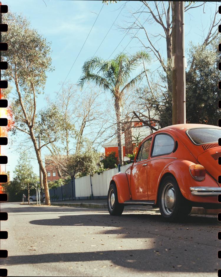 An orange volkswagen beetle is parked on a street next to a white fence and trees.