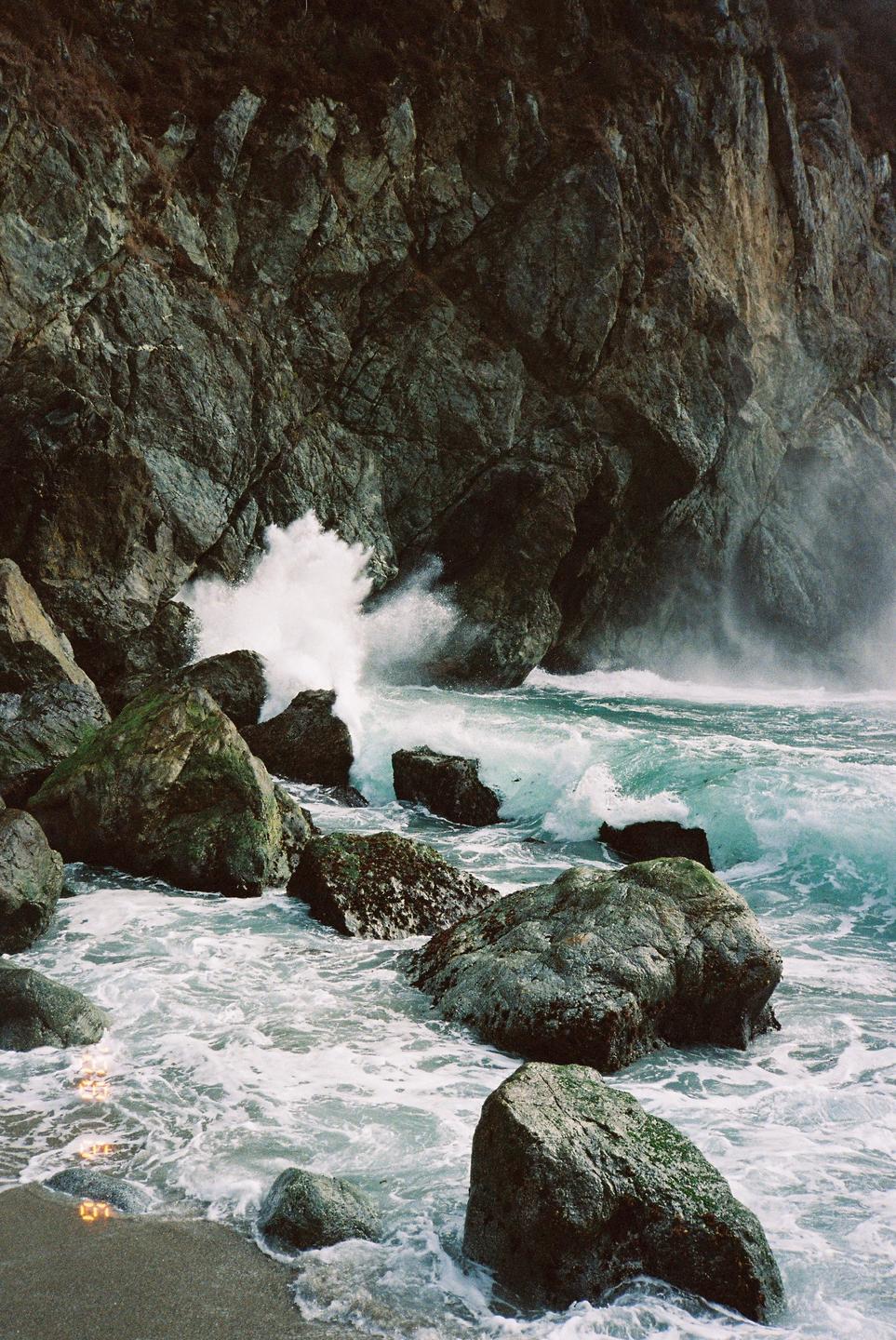 Waves crash against a rocky cliff and large boulders on a beach.
