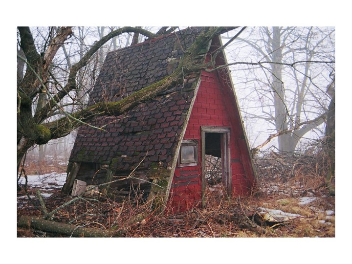 An abandoned red A-frame cabin with a dark roof stands in a winter forest.